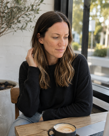 A woman with long brown hair sits at a wooden table in a cafe, wearing a black long-sleeve knit sweater and light blue jeans. A black mug with a latte is in the foreground, and a window with blurred outdoor scenery is behind her.